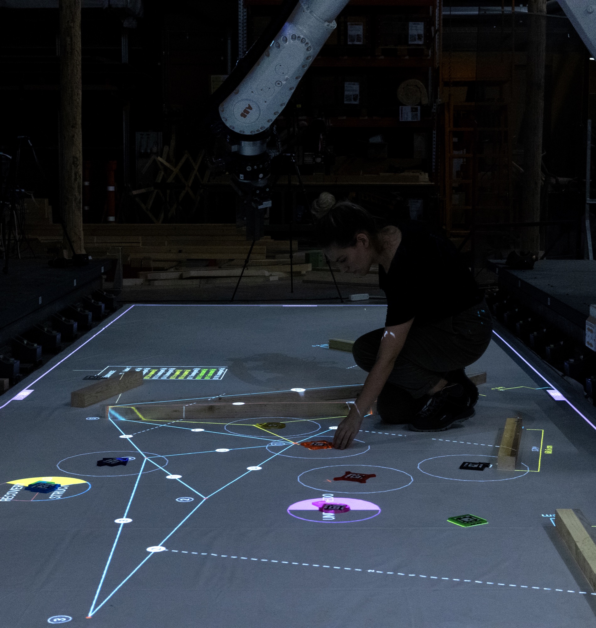 Person arranging timber offcuts on a floor projection of a truss design, beneath a robot arm